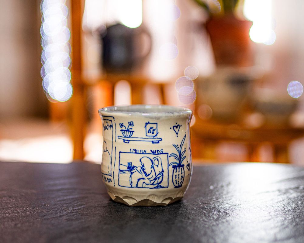 Handcrafted ceramic cup with blue illustrated scenes and text on dark stone surface with warm bokeh lighting.