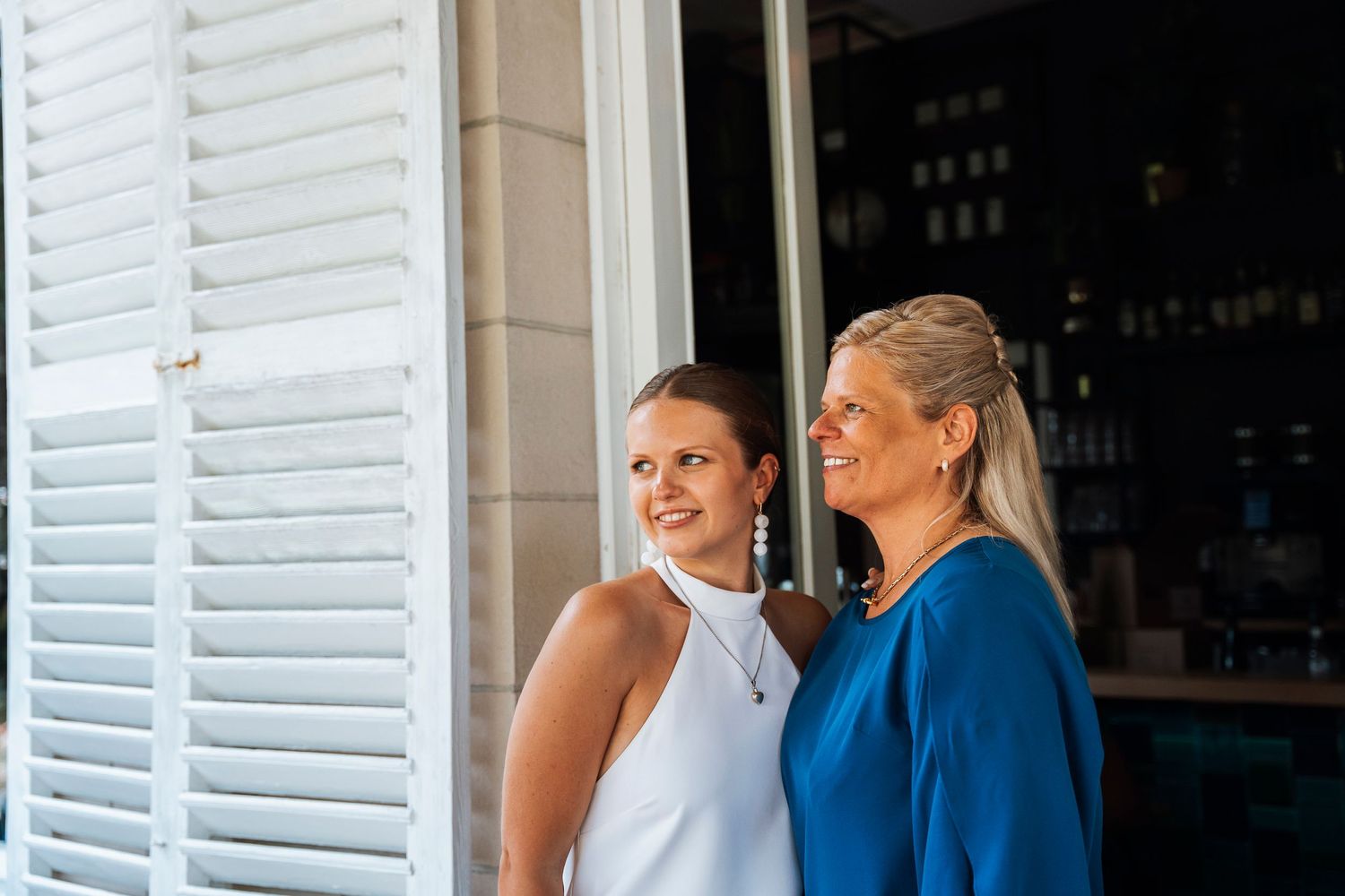 Two people in white and blue outfits smile together by white shutters.