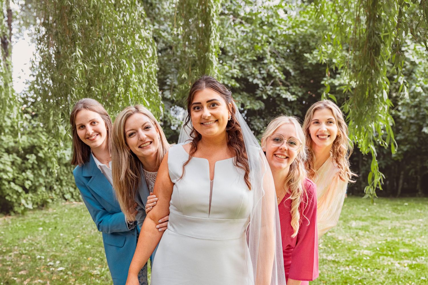 A group of wedding attendees pose playfully peeking from behind each other in a garden setting.