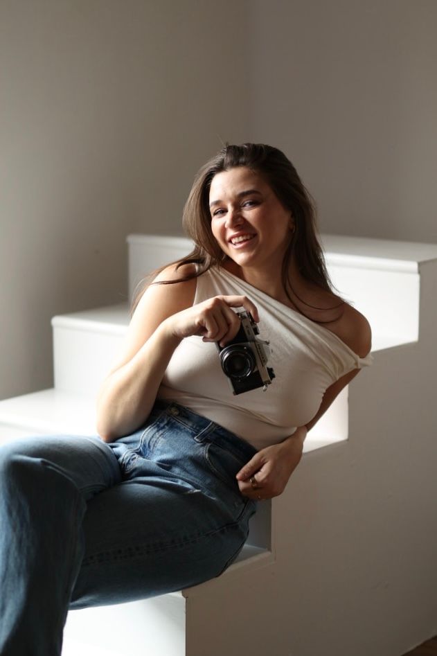 Smiling young woman sitting on white stairs holding a vintage film camera in casual attire.