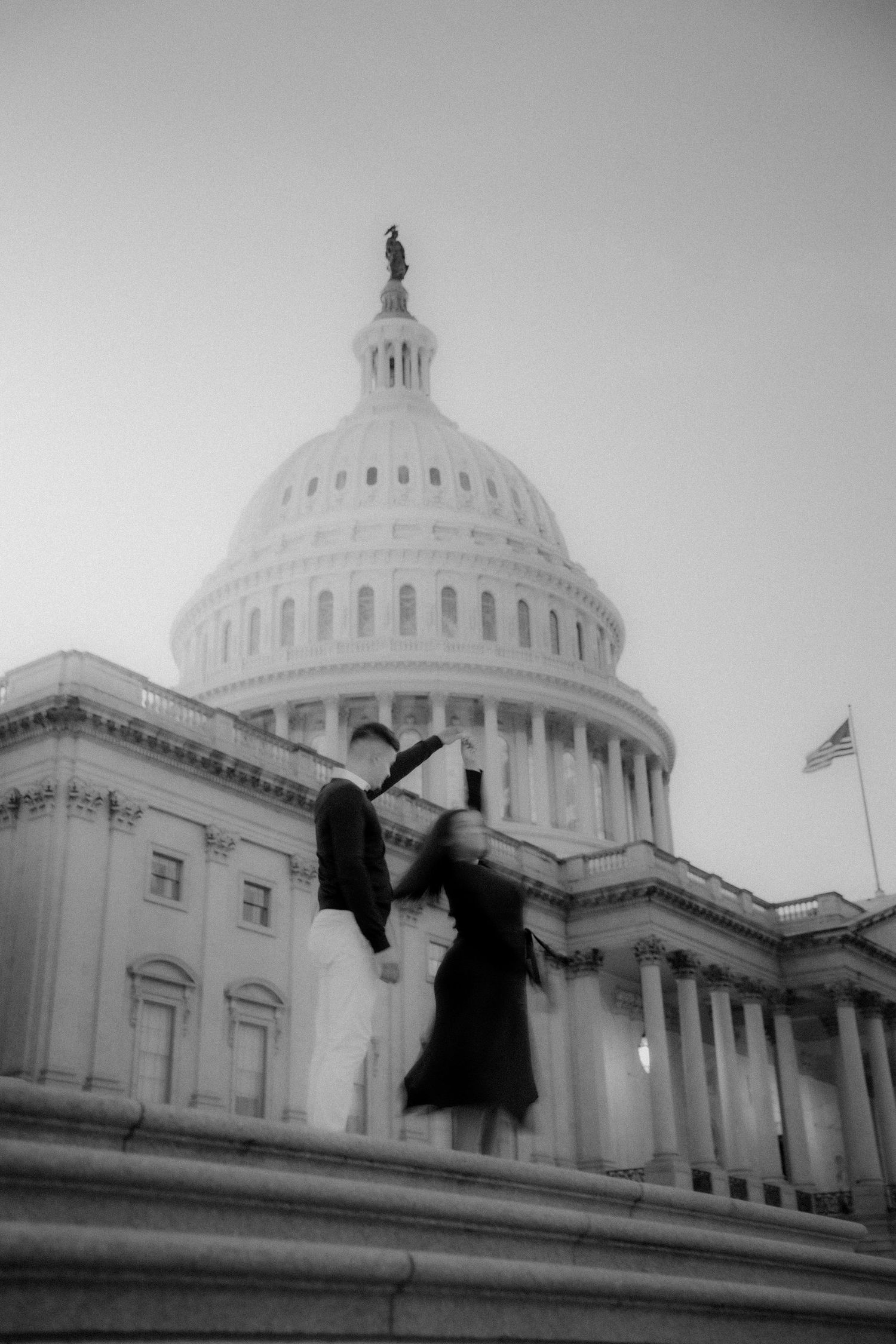 Two figures perform a dance pose against the backdrop of the U.S. Capitol Building dome in a black and white photograph.
