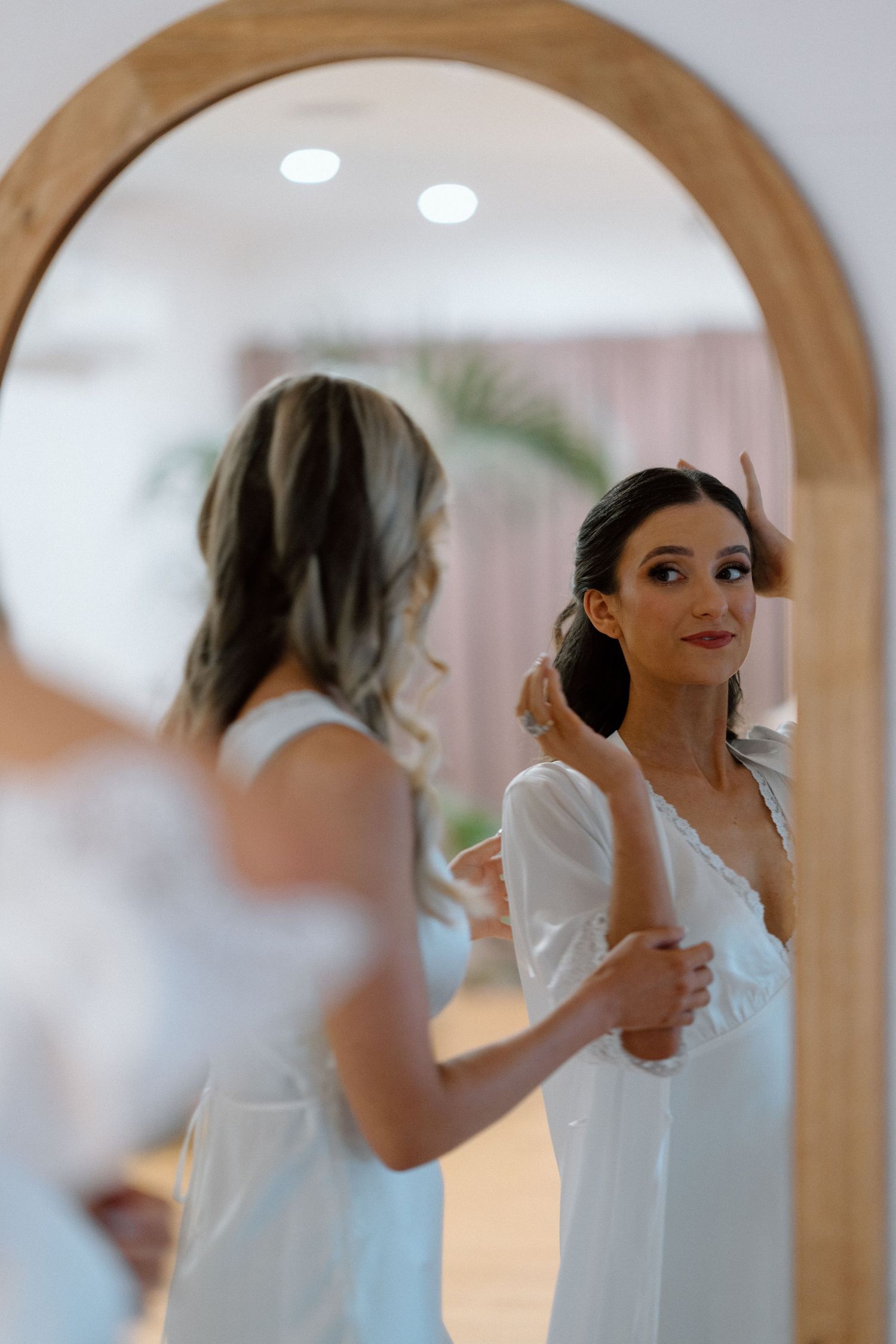 Reflection of bride getting ready in round wooden mirror showing delicate white gown details.