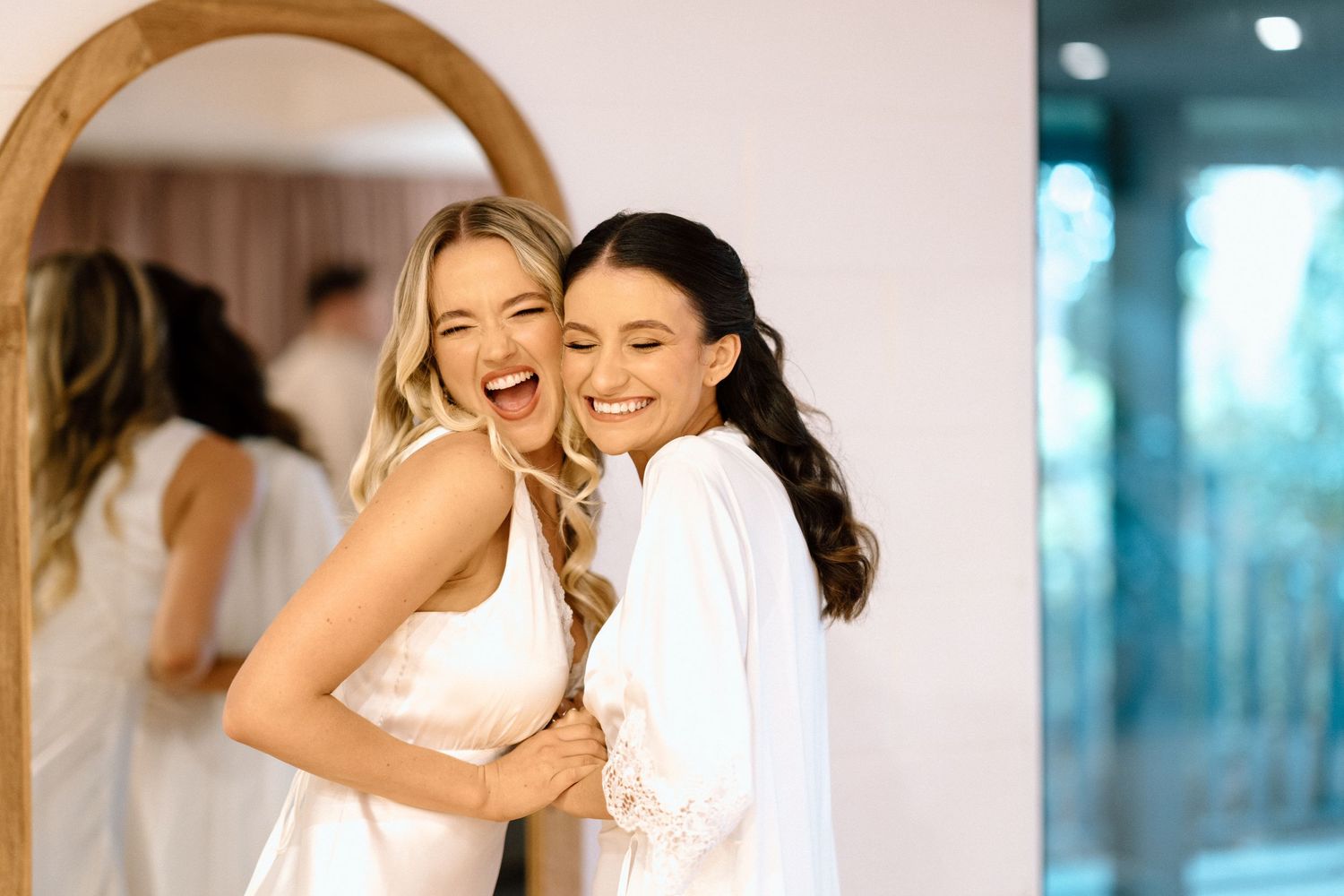 Two women in white robes sharing joyful laughter while getting ready in dressing room.