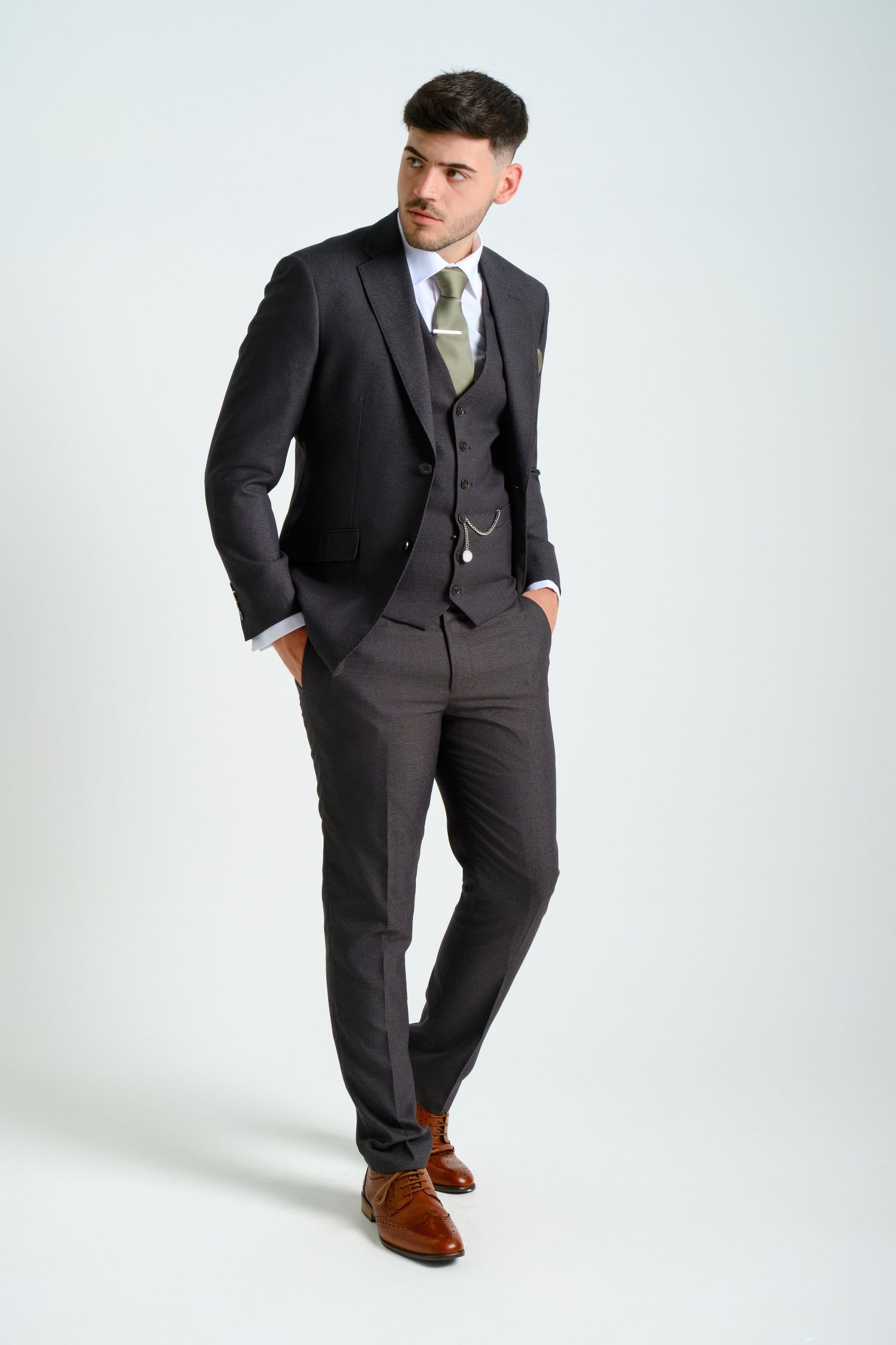 Male model in tailored dark suit with olive tie, posing confidently with hands in pockets against a plain background