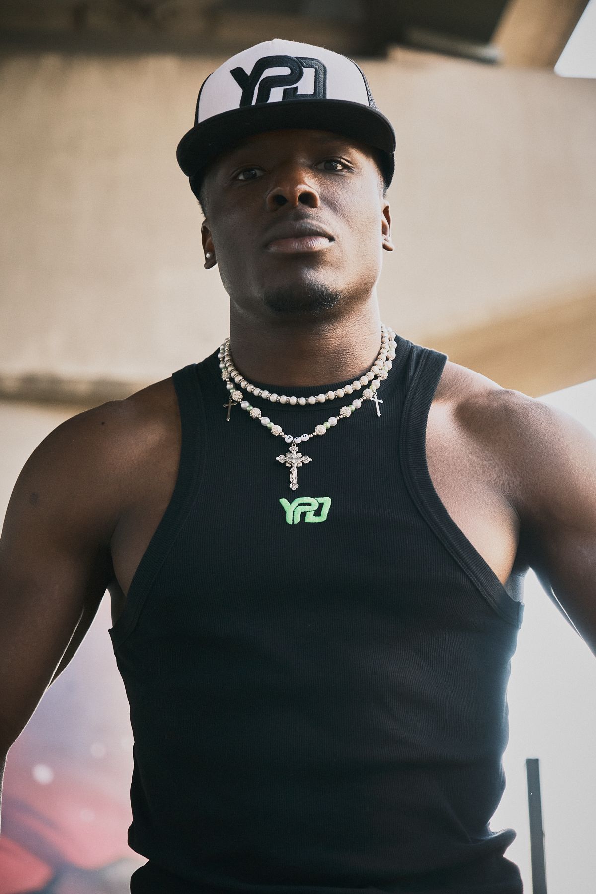 Male model in black tank top, layered cross necklaces, and a cap, creating a streetwear look under urban lighting