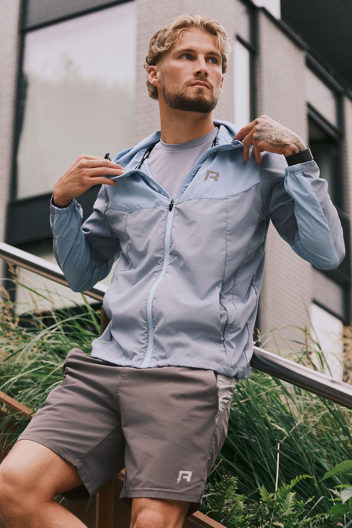 Male model in blue athletic jacket and grey shorts, adjusting collar while posing outdoors near urban architecture