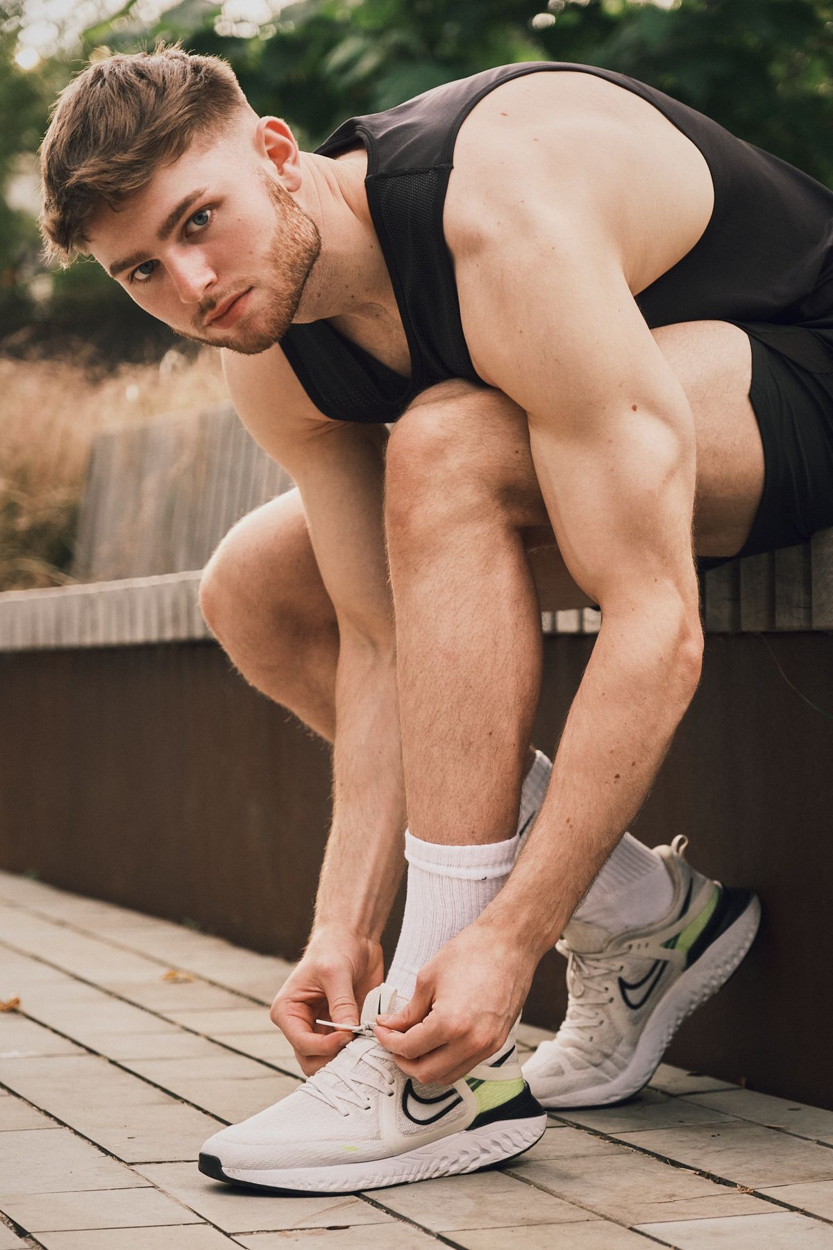 Male model in black tank top, lacing white trainers, creating a sporty, casual look with focus on footwear.