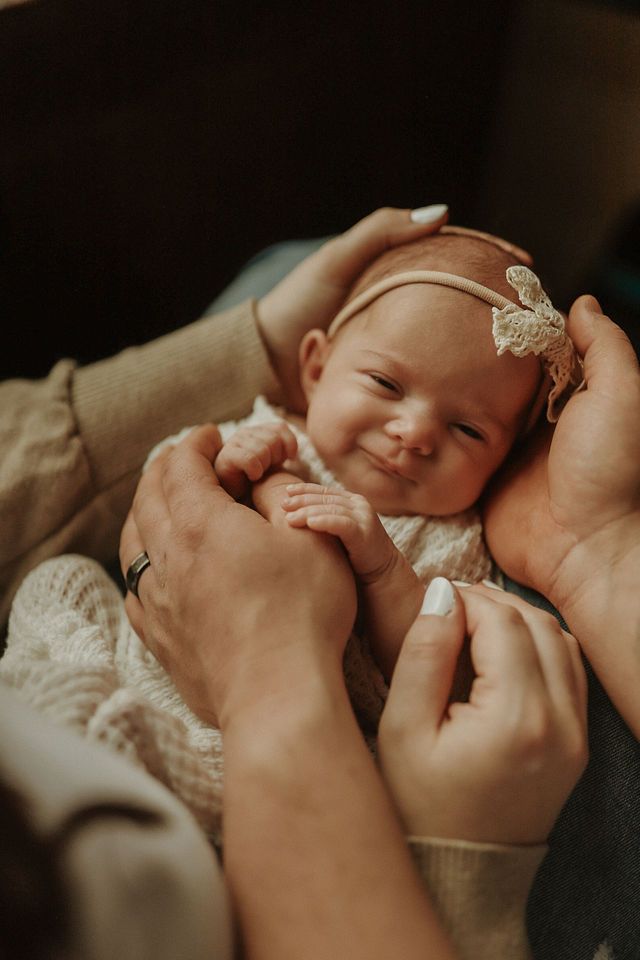 A newborn baby wearing a delicate headband smiles while being cradled in loving hands.