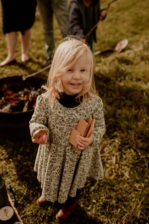 Young child in floral dress smiles while holding marshmallow stick near campfire.