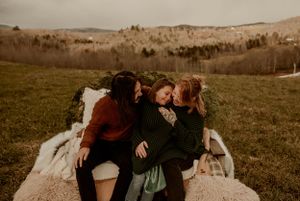 Three friends share an intimate moment sitting together on a blanket in a grassy field at sunset.