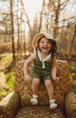 Joyful child in overalls and cap laughs while sitting on vintage patterned armchair outdoors.