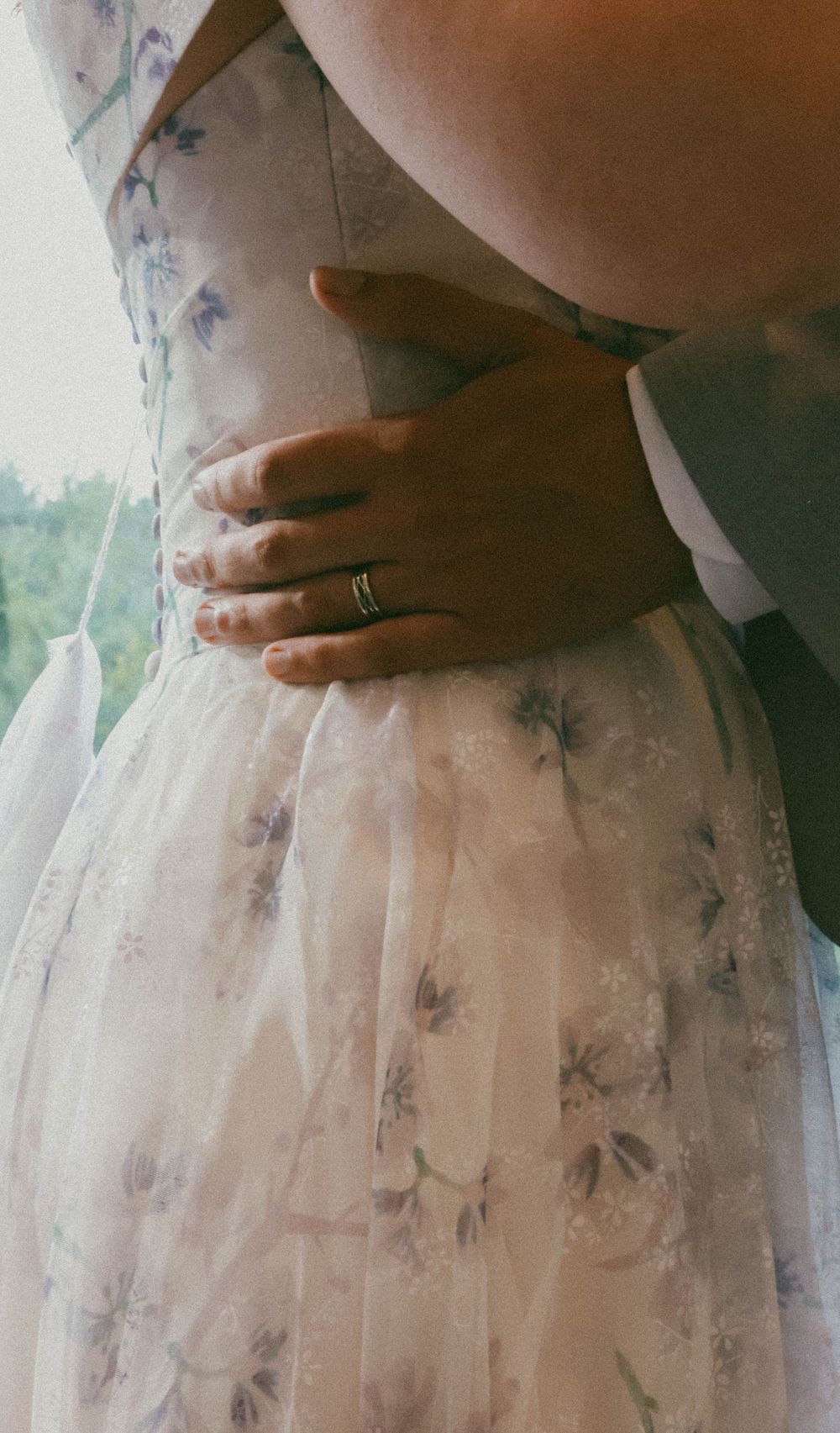 Close-up of groom's hand with gold wedding band embracing bride in floral gown.