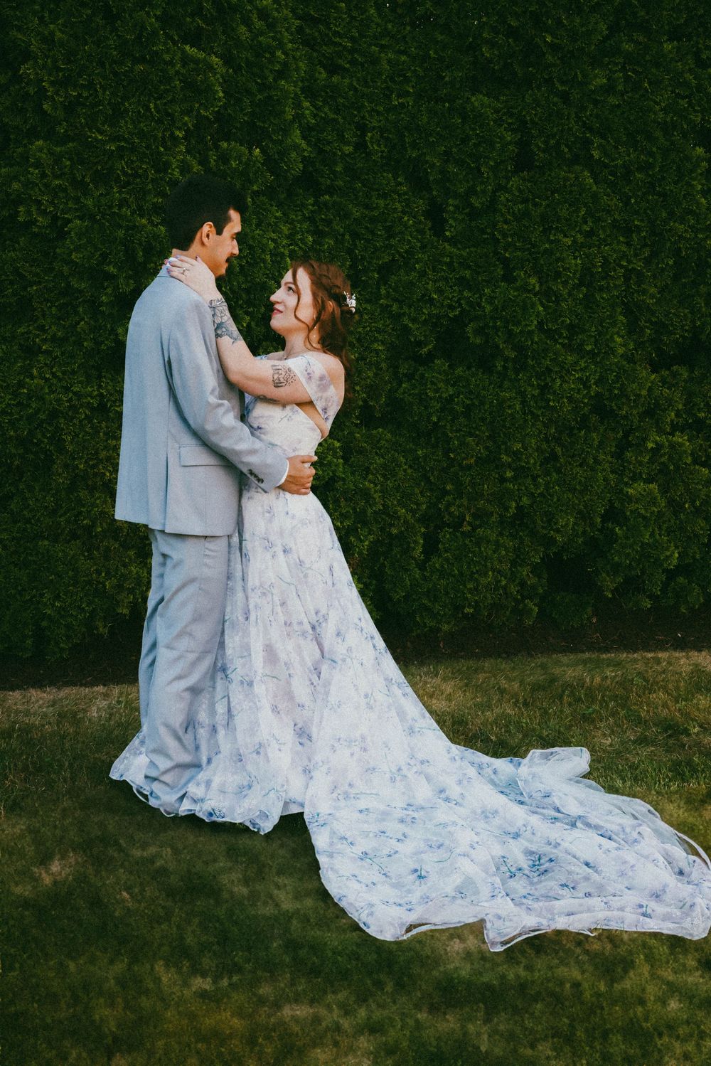 Bride in floral blue and white gown embracing groom in gray suit against green hedge.