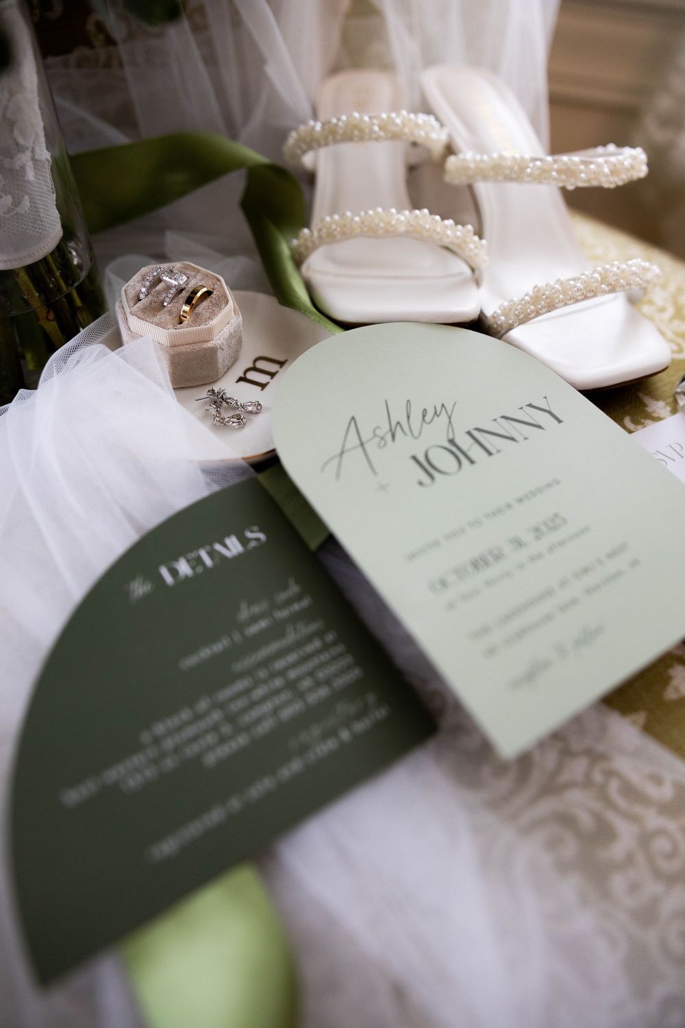 Wedding flat lay featuring an invitation for Ashley and Johnny, pearl heels, rings, and a veil.