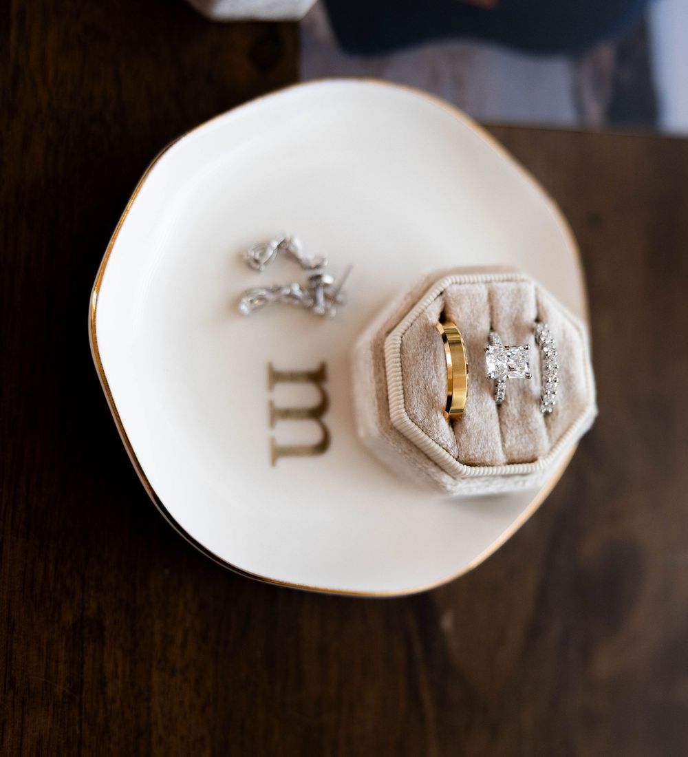 A gold wedding band and diamond engagement ring displayed in a velvet ring box on a ceramic dish.