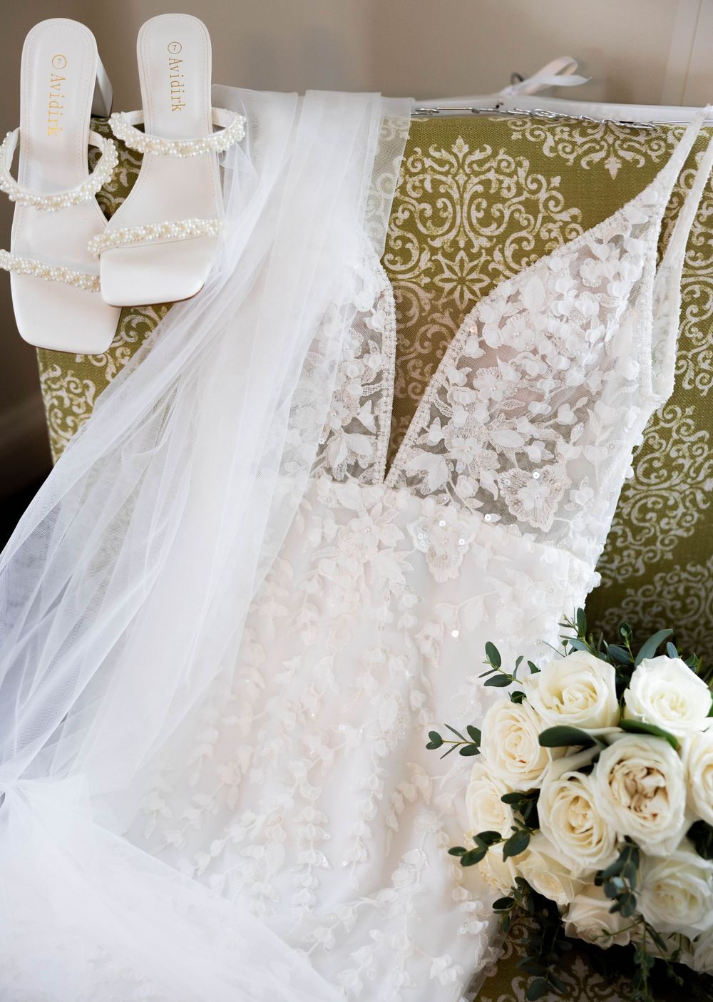Flat lay of lace wedding dress, white pearl heels, veil, and white rose bouquet.