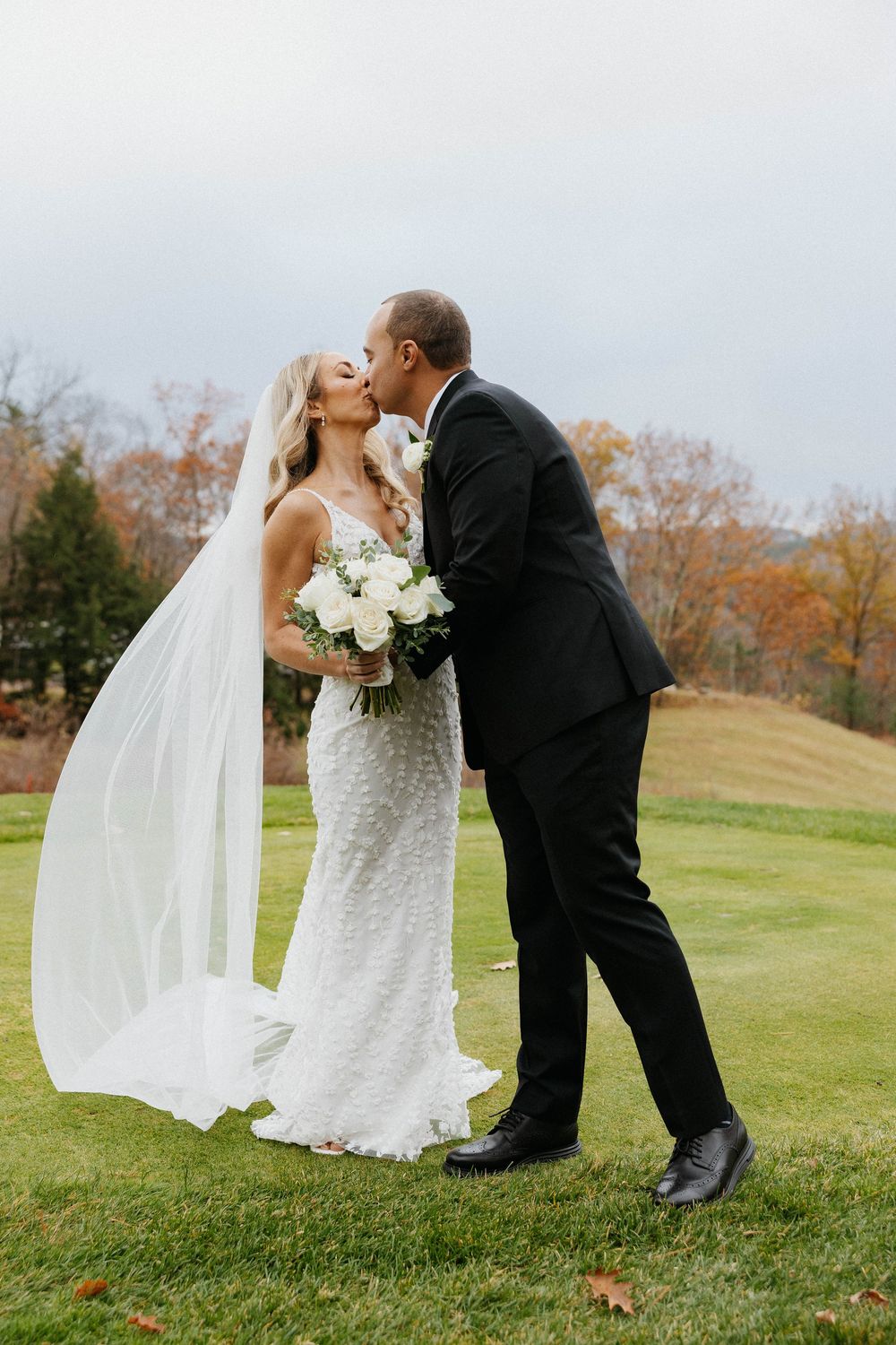 A bride and groom share a kiss outdoors on a green lawn with autumn foliage in the background.