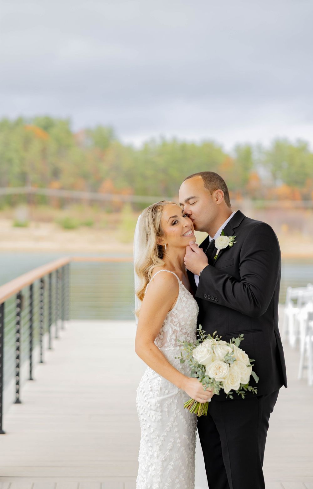 A bride and groom share a tender moment on a waterfront dock, bride holding a white rose bouquet.