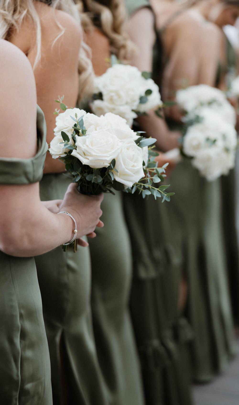 A row of bridesmaids in sage green dresses holding white rose and eucalyptus bouquets.