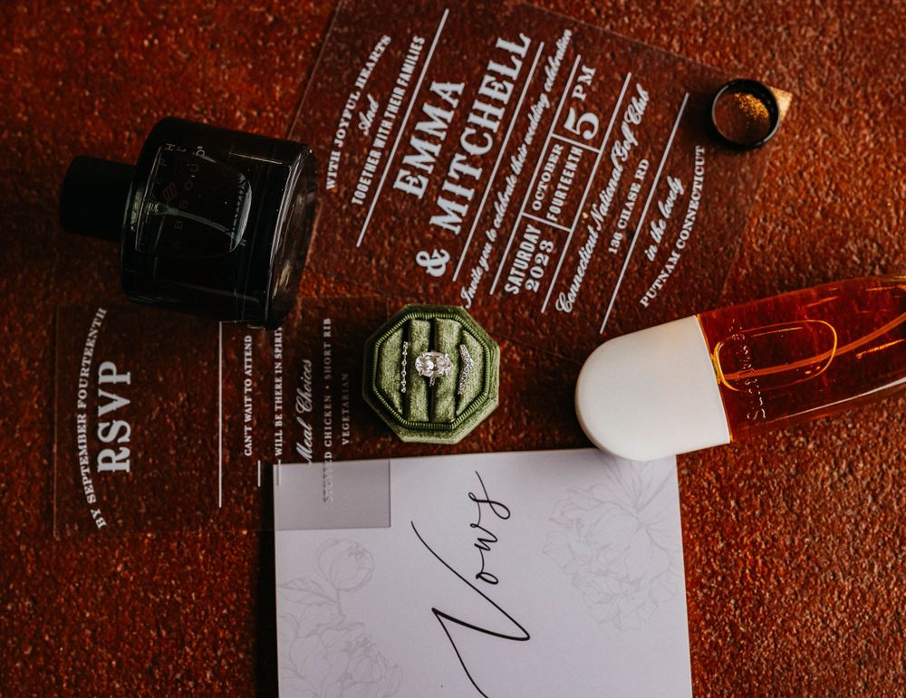Wedding invitation suite with acrylic invite, wax seal, ring, and perfume bottle on rustic background.