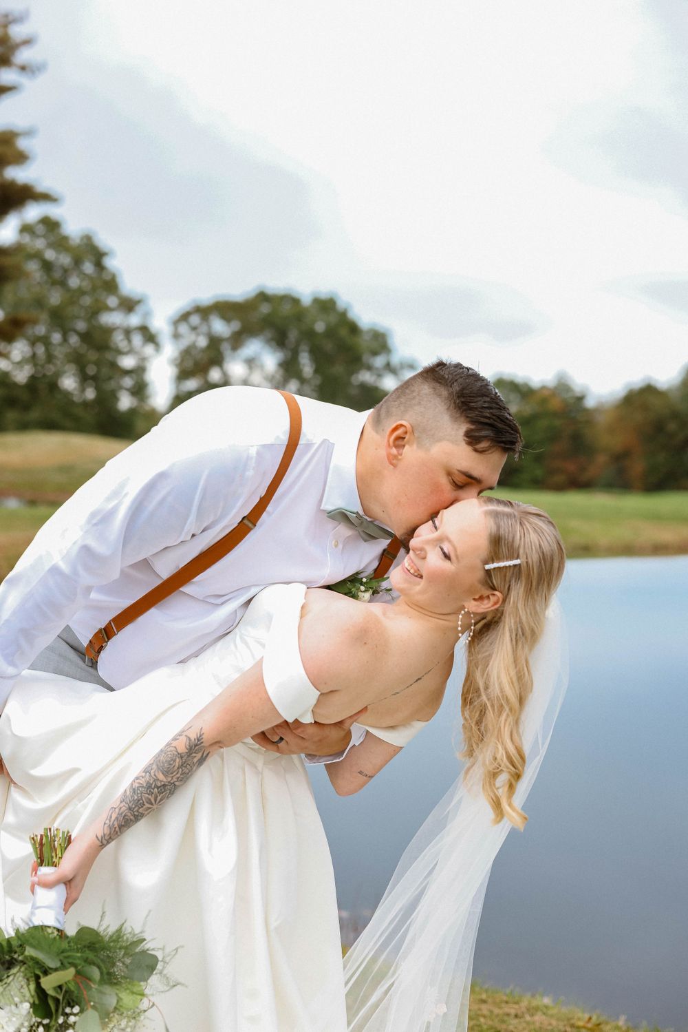 Groom dips and kisses bride in white gown with veil beside a serene pond outdoors.