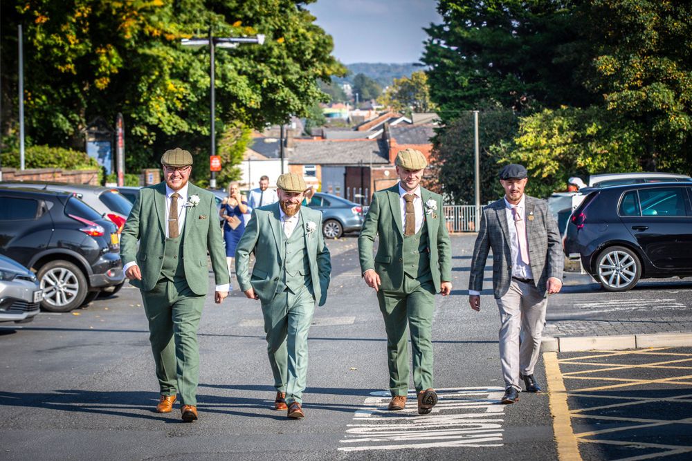 Four groomsmen in matching sage green suits walking confidently through a sunny car park.