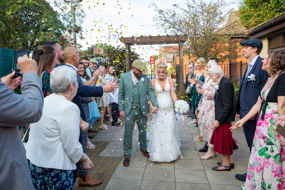 Smiling bride and groom walk through a confetti shower surrounded by cheering guests outside a venue.