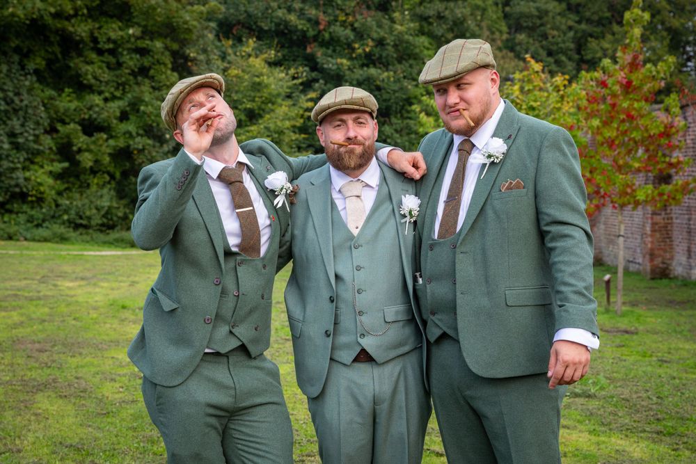 Three groomsmen in sage green suits and flat caps share a fun candid moment outdoors.