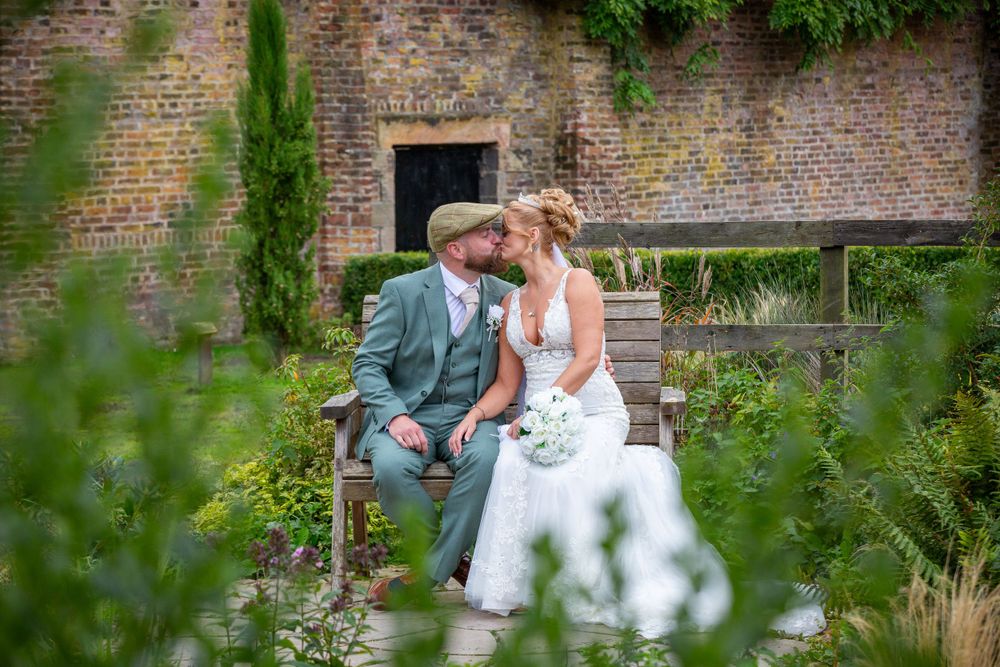 Bride and groom share a romantic kiss on a garden bench surrounded by lush greenery and a rustic brick wall.