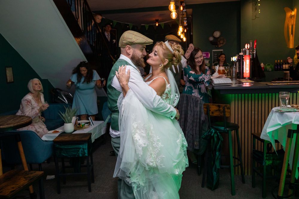 Bride and groom share a joyful first dance embrace in a vibrant green bar venue with wedding guests celebrating.