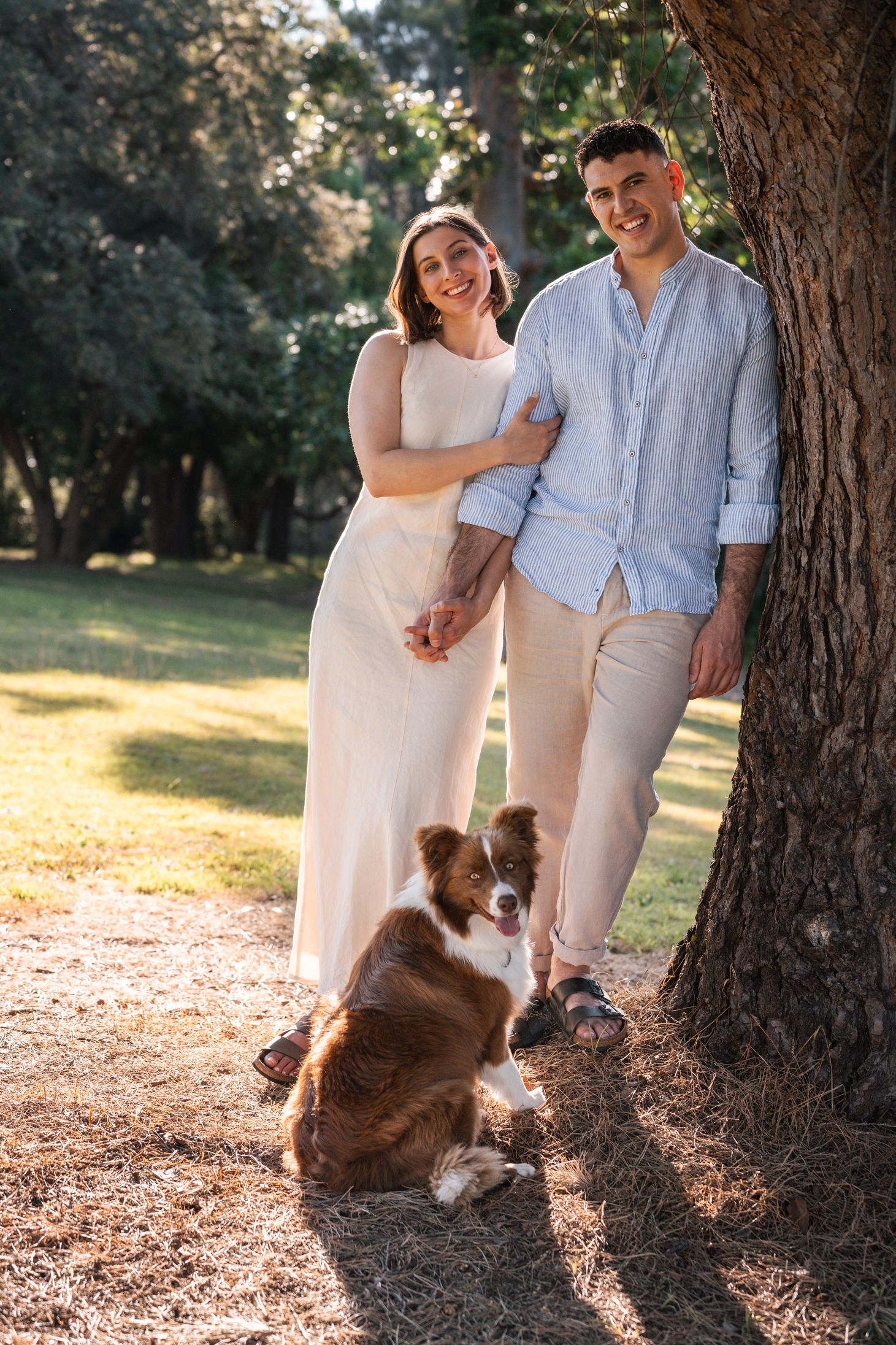 man-leaning-on-the-tree-and-smiling-while-women-hugs-him-and-smiles-too-together-with-their-border-collie-during-a-coupl