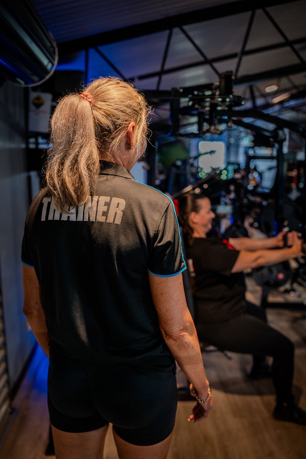 A personal trainer in a black uniform watches a client exercising at a dimly lit modern gym facility.