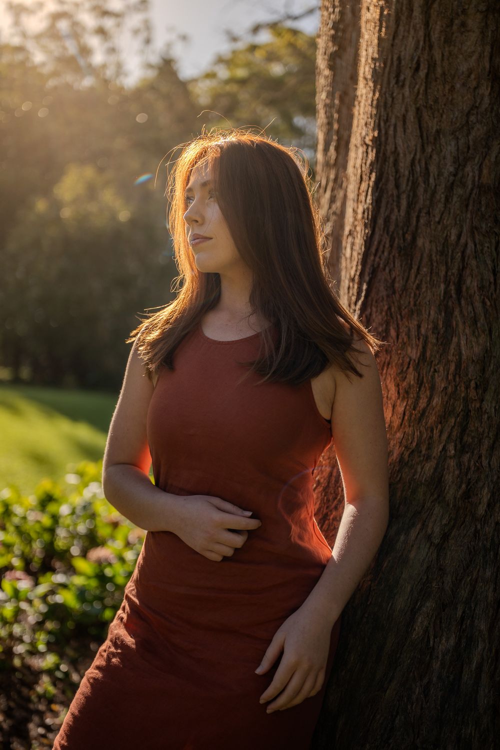 A woman in a burgundy dress leans against a tree trunk at sunset, golden light illuminating the peaceful nature scene.