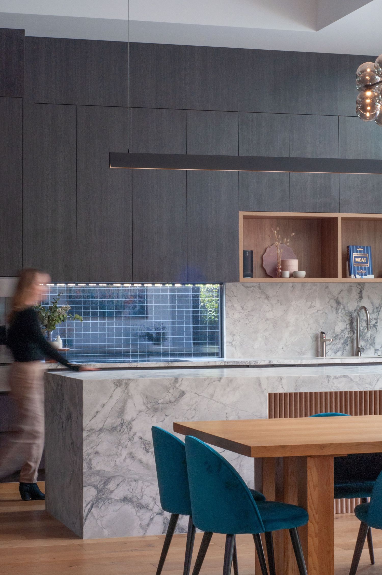 Modern kitchen interior with marble countertops, dark wall panels, and teal dining chairs.