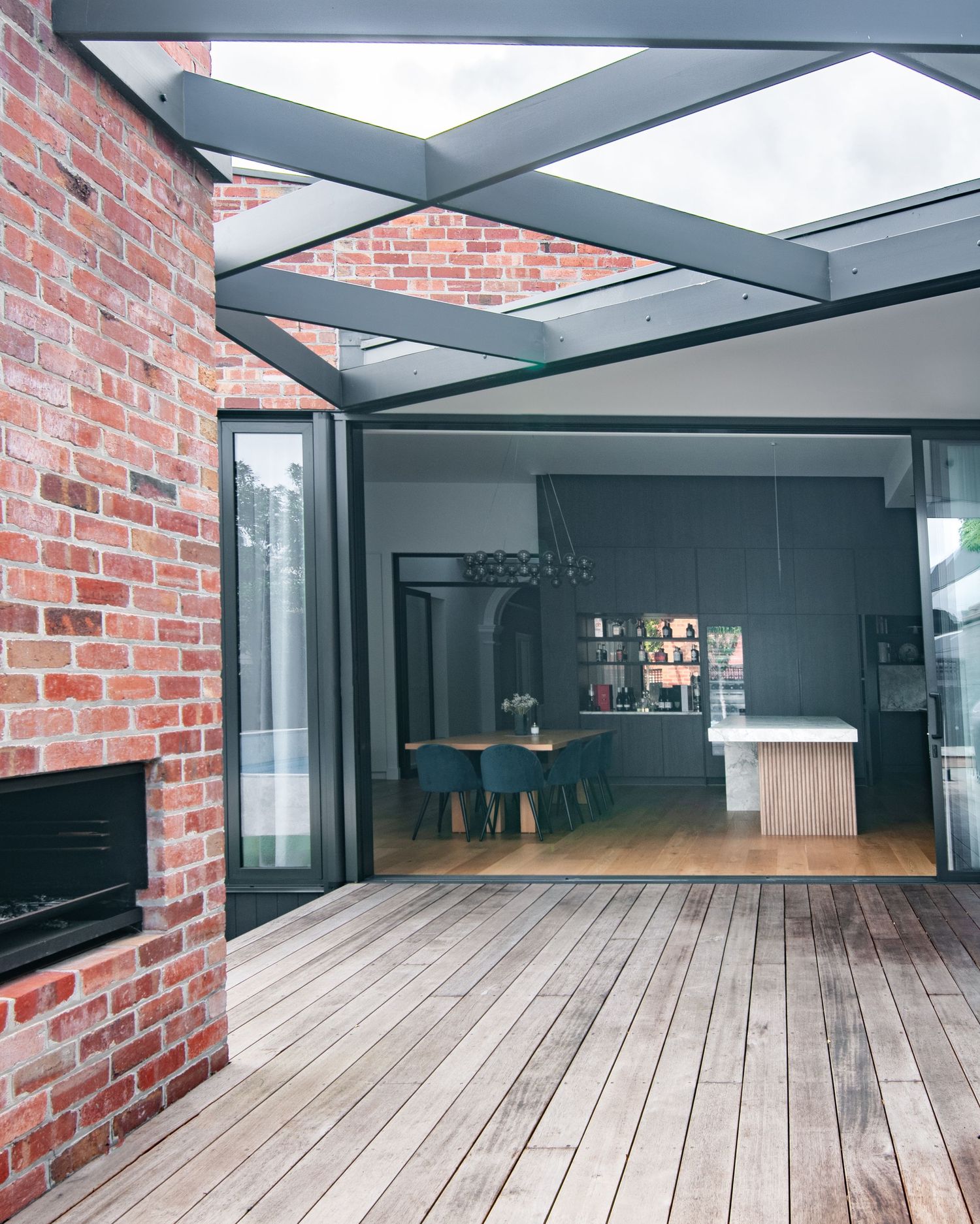 Modern indoor-outdoor living space with exposed brick wall, glass ceiling panels, and wooden deck flooring.