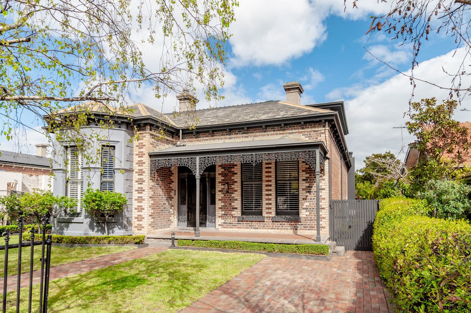 Historic stone cottage with wrap-around veranda and manicured garden behind iron fence.