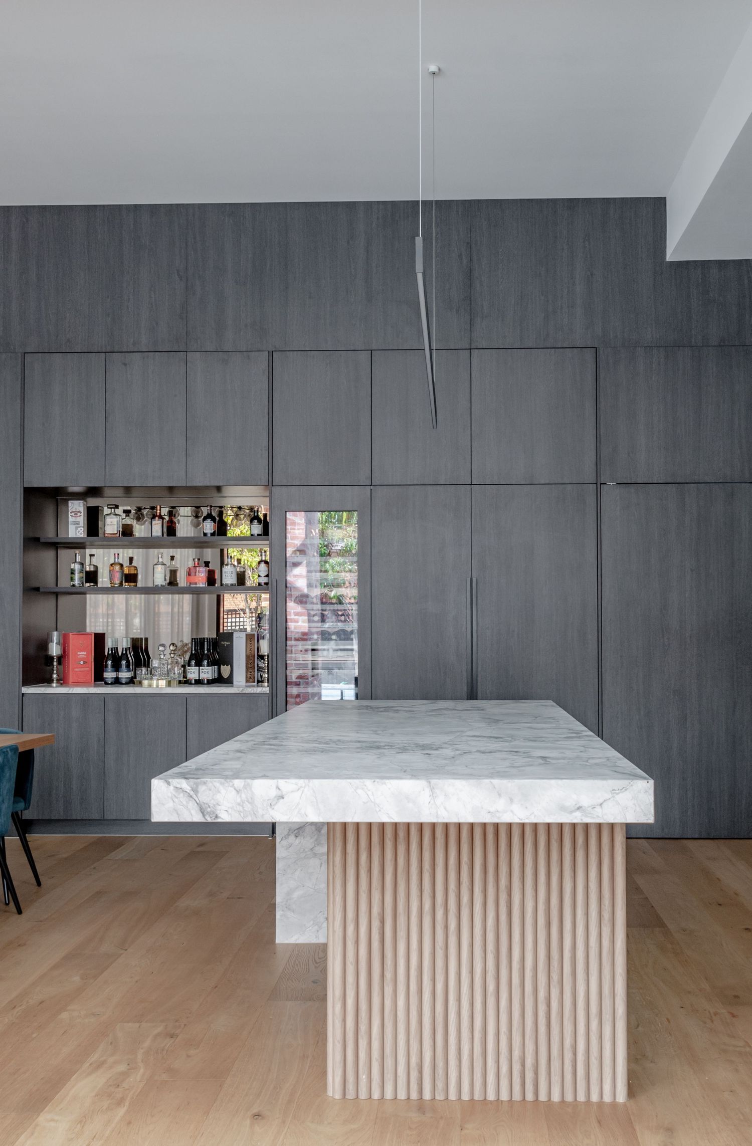 Luxurious kitchen island with marble top and wooden slat base against dark paneled walls with built-in bar.