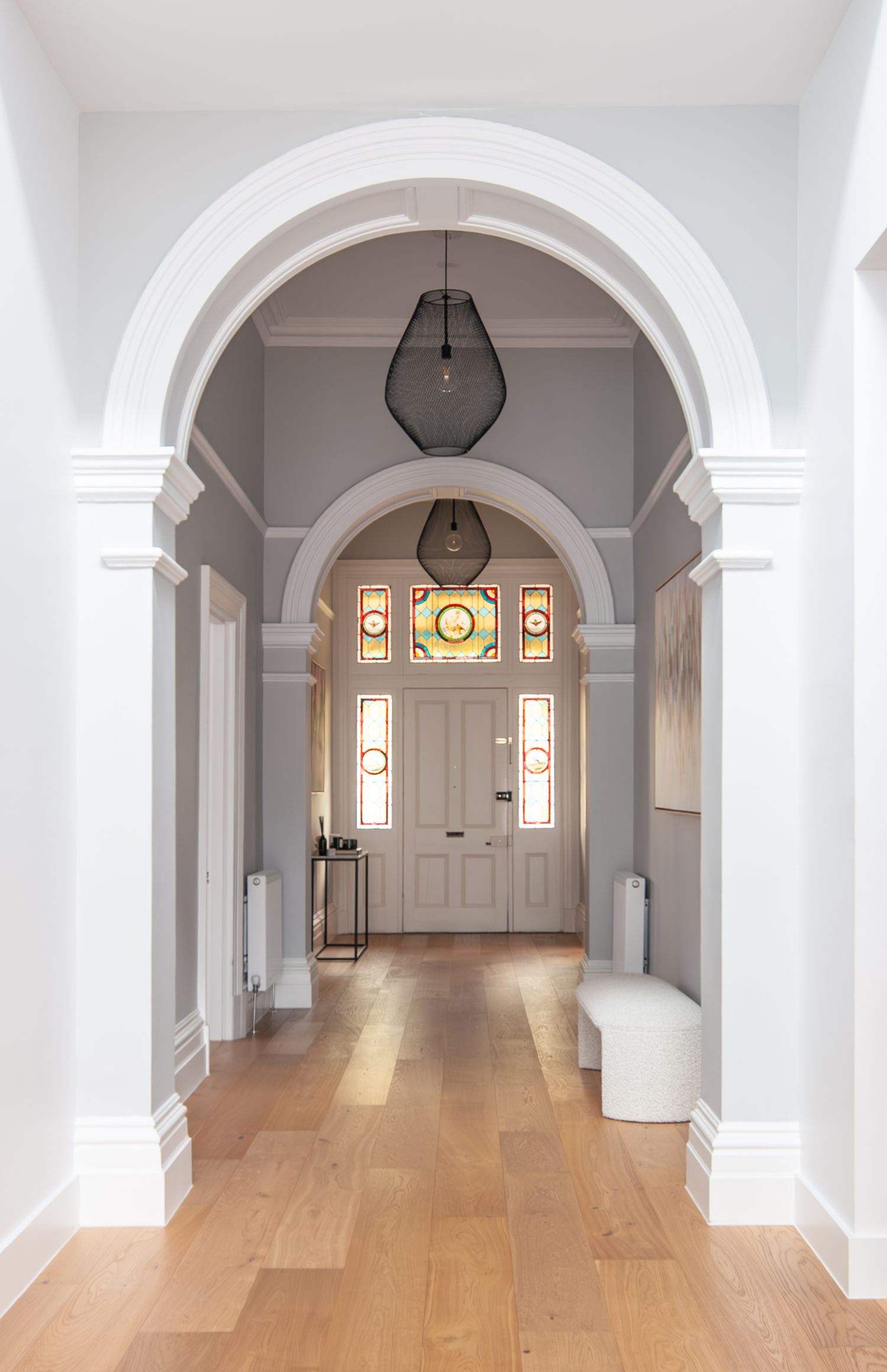 Classical hallway with arched doorways, hardwood floors, and pendant lighting leading to a stained glass front door.