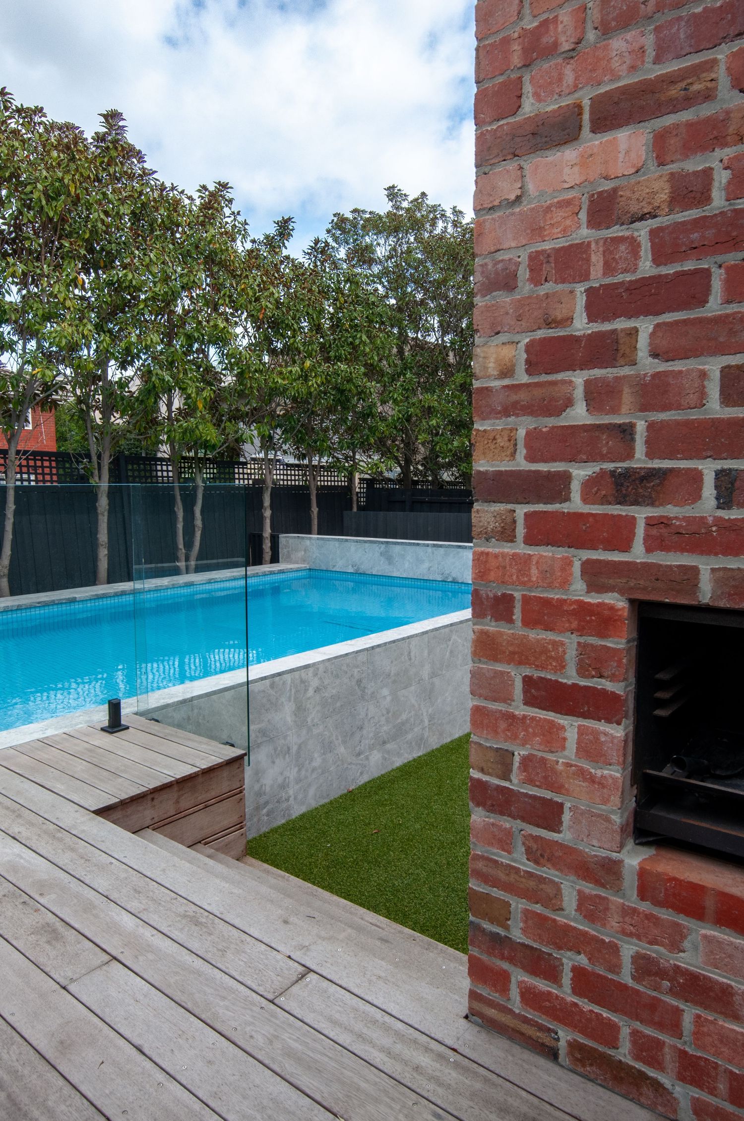 Outdoor pool area with brick fireplace, wooden decking, and glass safety barriers.