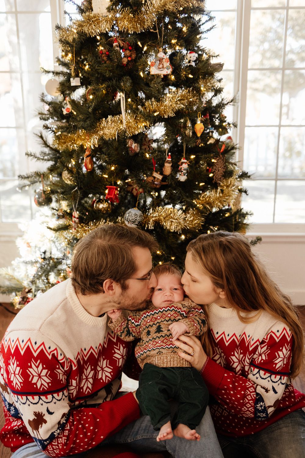 Parents kiss baby's cheeks in front of Christmas tree in San Jose at in-home newborn session with Bay Area Photographer