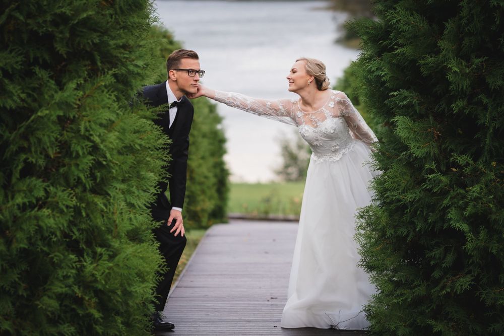 Playful wedding couple poses on a wooden boardwalk surrounded by green hedges.