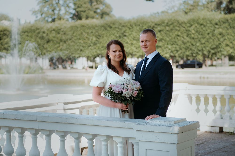 A couple stands on a white stone balcony with ornate railings near a fountain, with green foliage in the background.