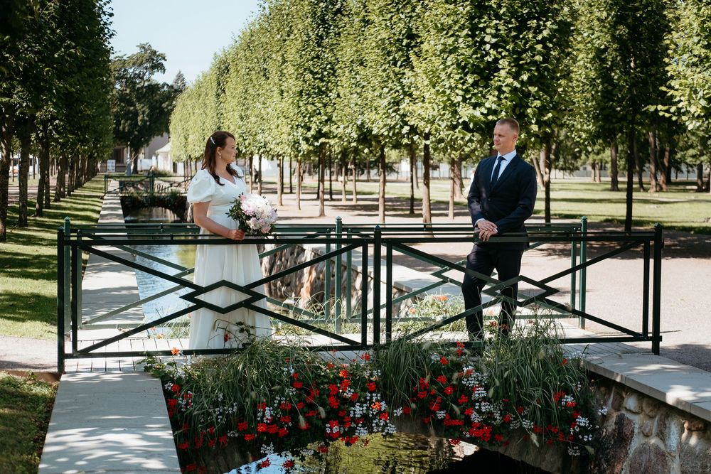 Two people stand on opposite sides of a decorative metal railing in a manicured garden path lined with trees and red flowers.