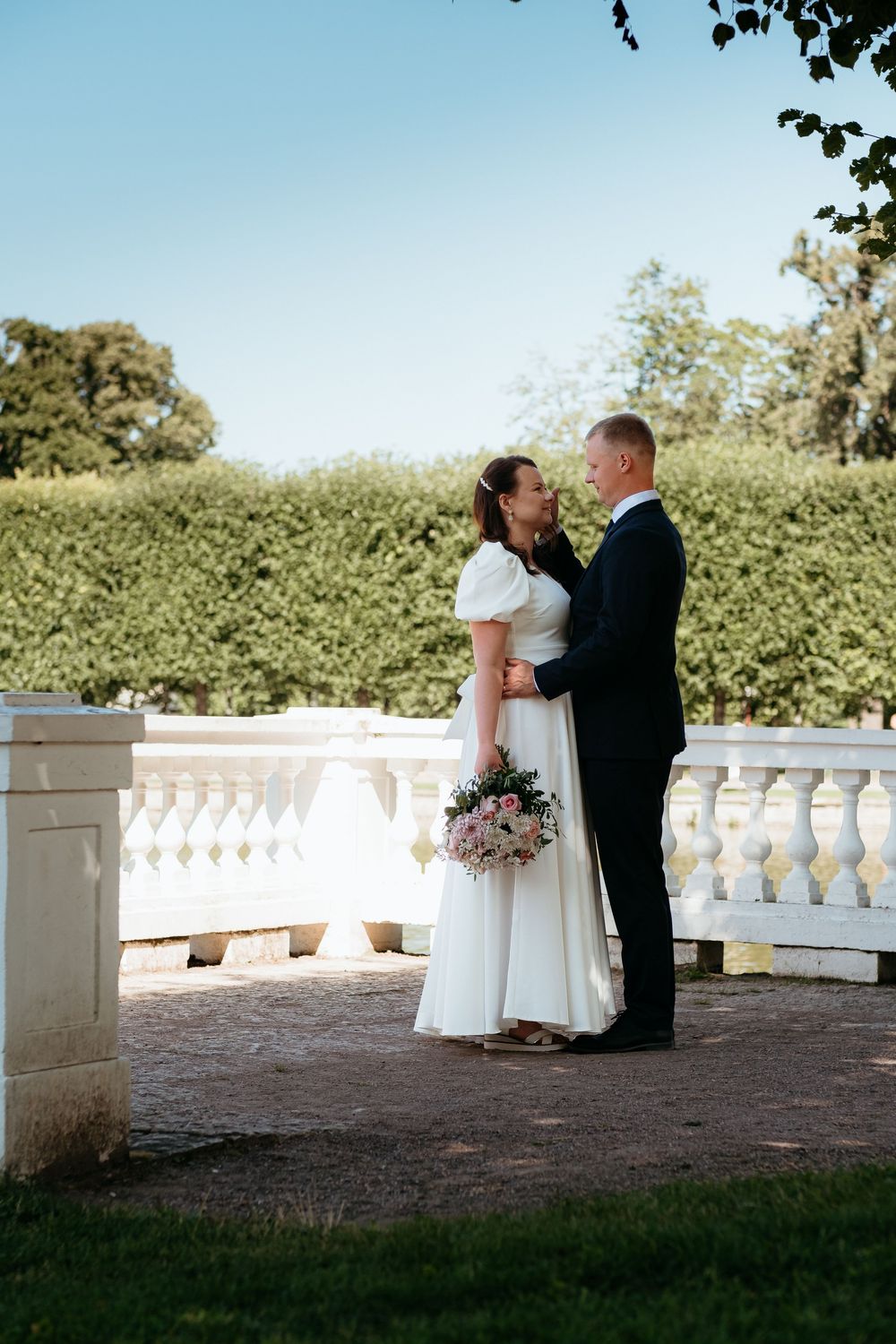 A romantic wedding couple embraces on an elegant white stone terrace surrounded by lush greenery on a sunny day.