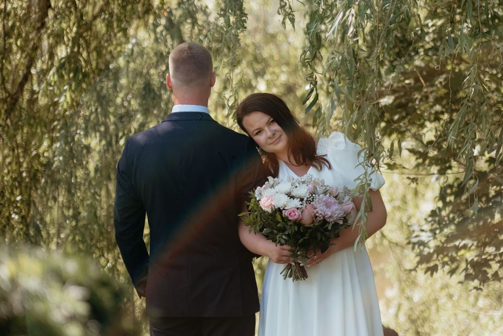 A couple in wedding attire shares a tender moment under willow trees in a romantic outdoor setting.