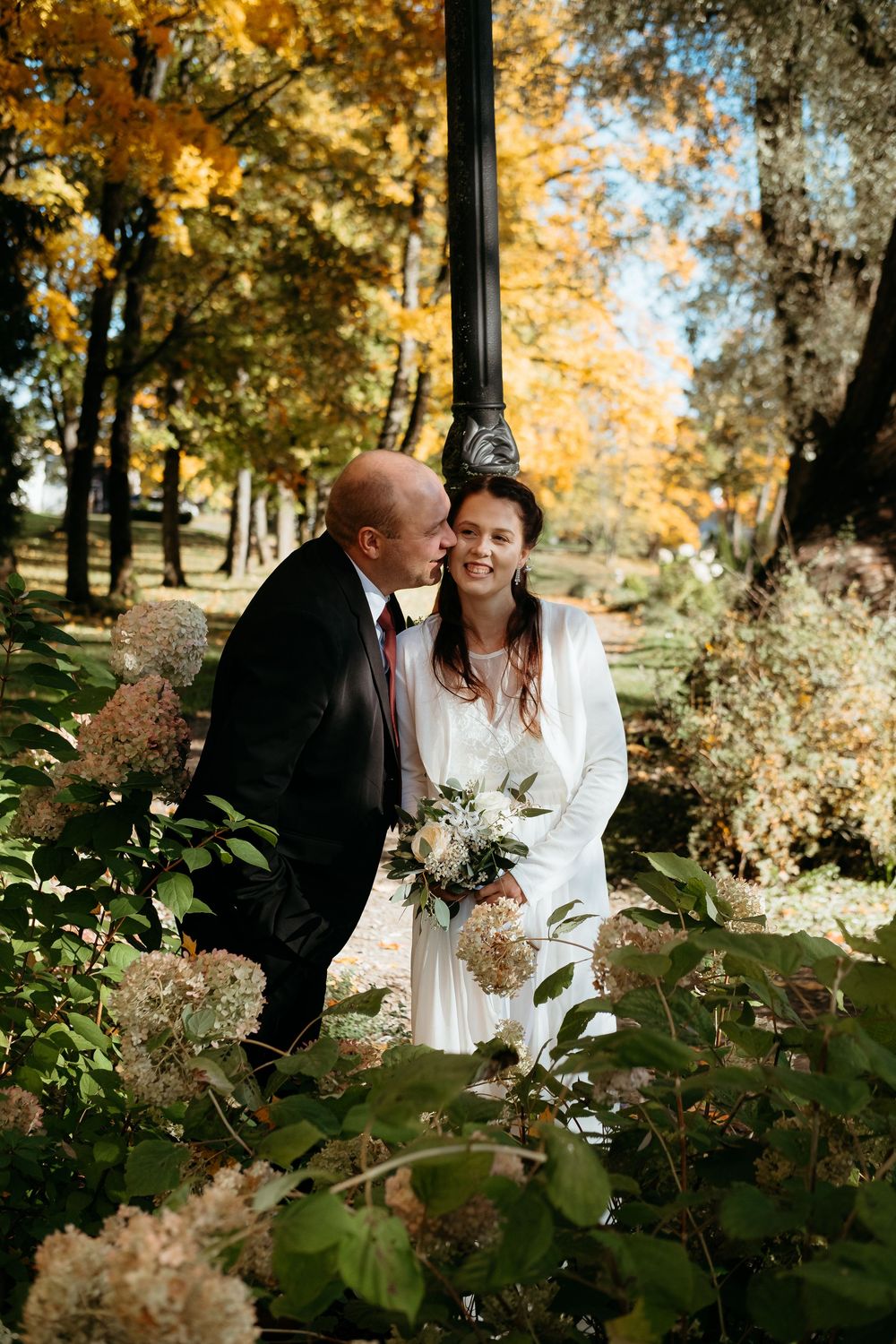 A romantic couple shares intimate moments in a sun-dappled autumn garden setting with golden foliage in the background.