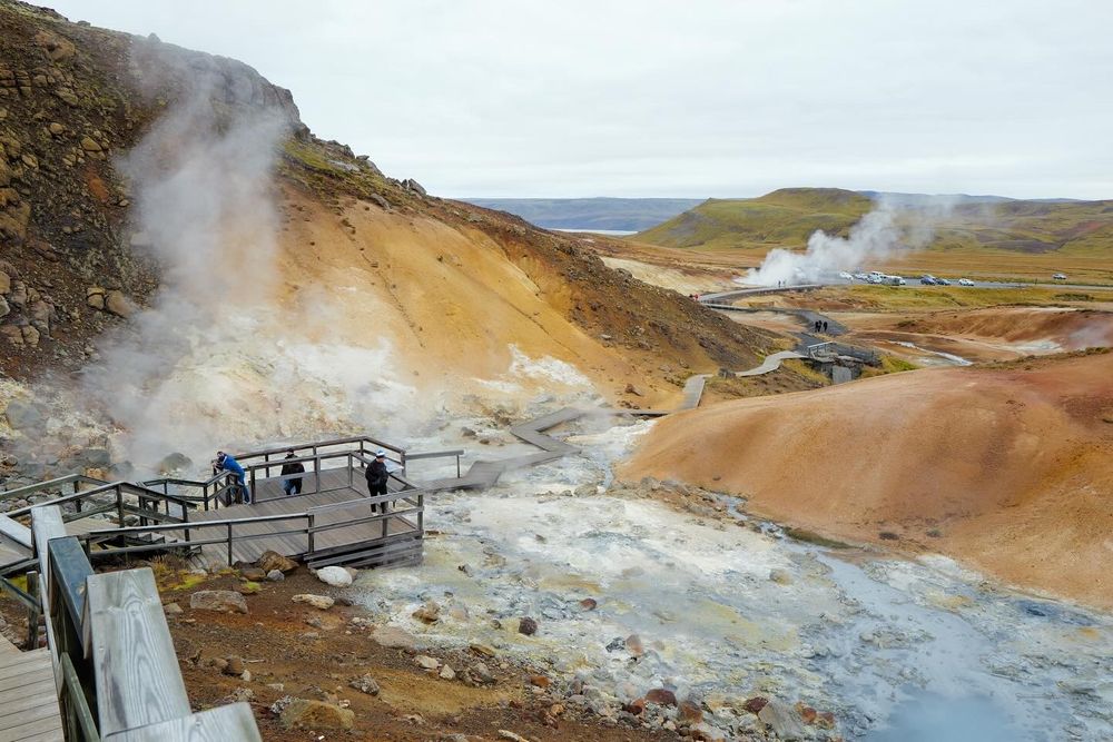 Steaming geothermal area with viewing platform and colorful mineral deposits in volcanic terrain.