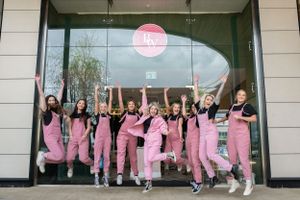 A group wearing matching pink jumpsuits jumps excitedly in front of a storefront with a pink circular sign.