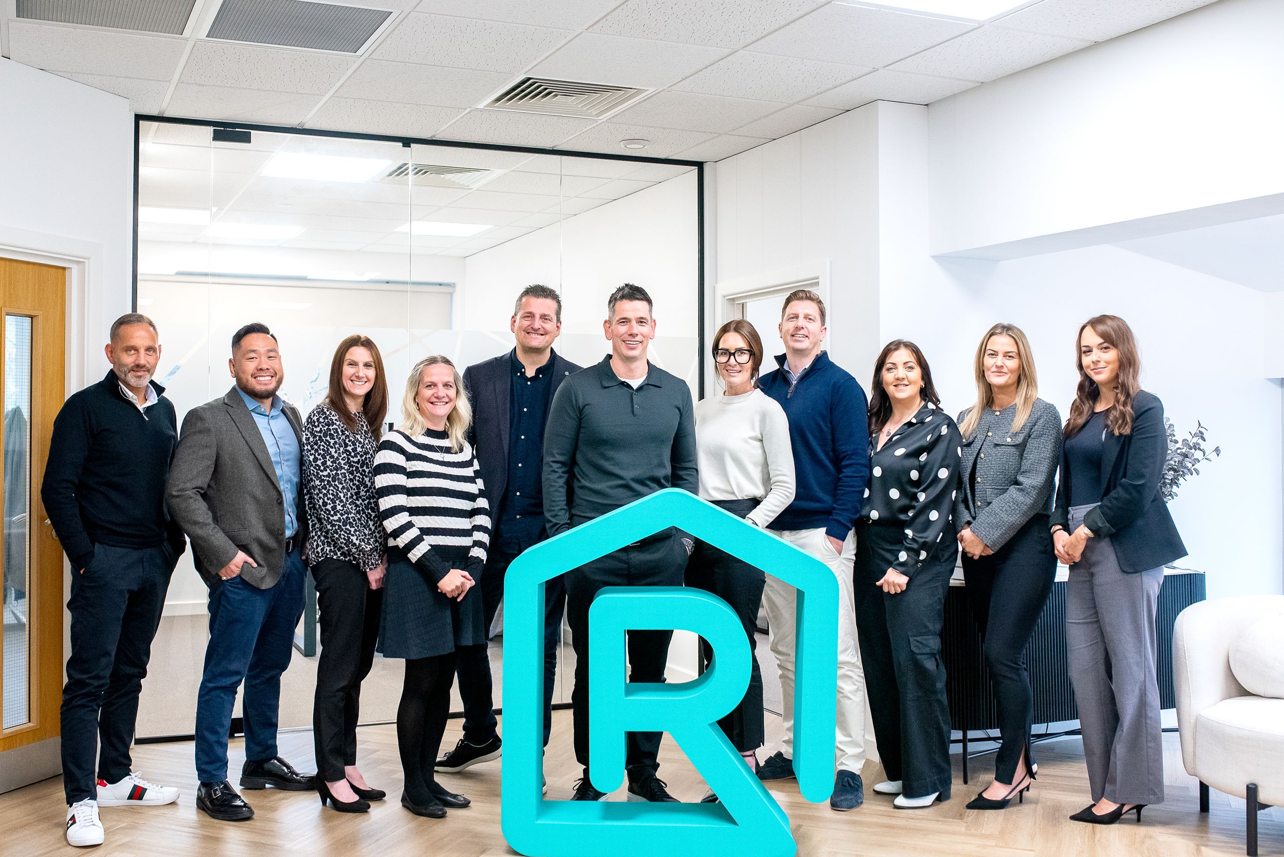 A group of professionals stand together in an office with a large turquoise R logo displayed in front of them.