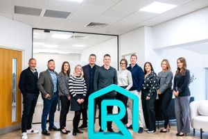 Group photo of business professionals standing together in a modern office space with a bright blue logo display.